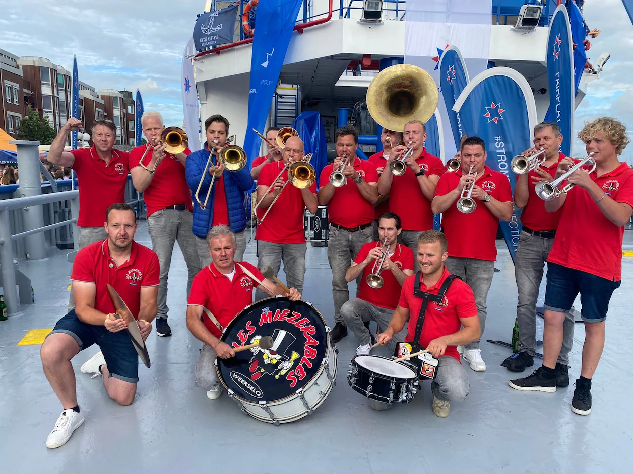 Recente kleurenfoto: de huidige Les Miezerables in rode shirts met blaasinstrumenten en de basdrum 'Les Miezerables Weerselo'.